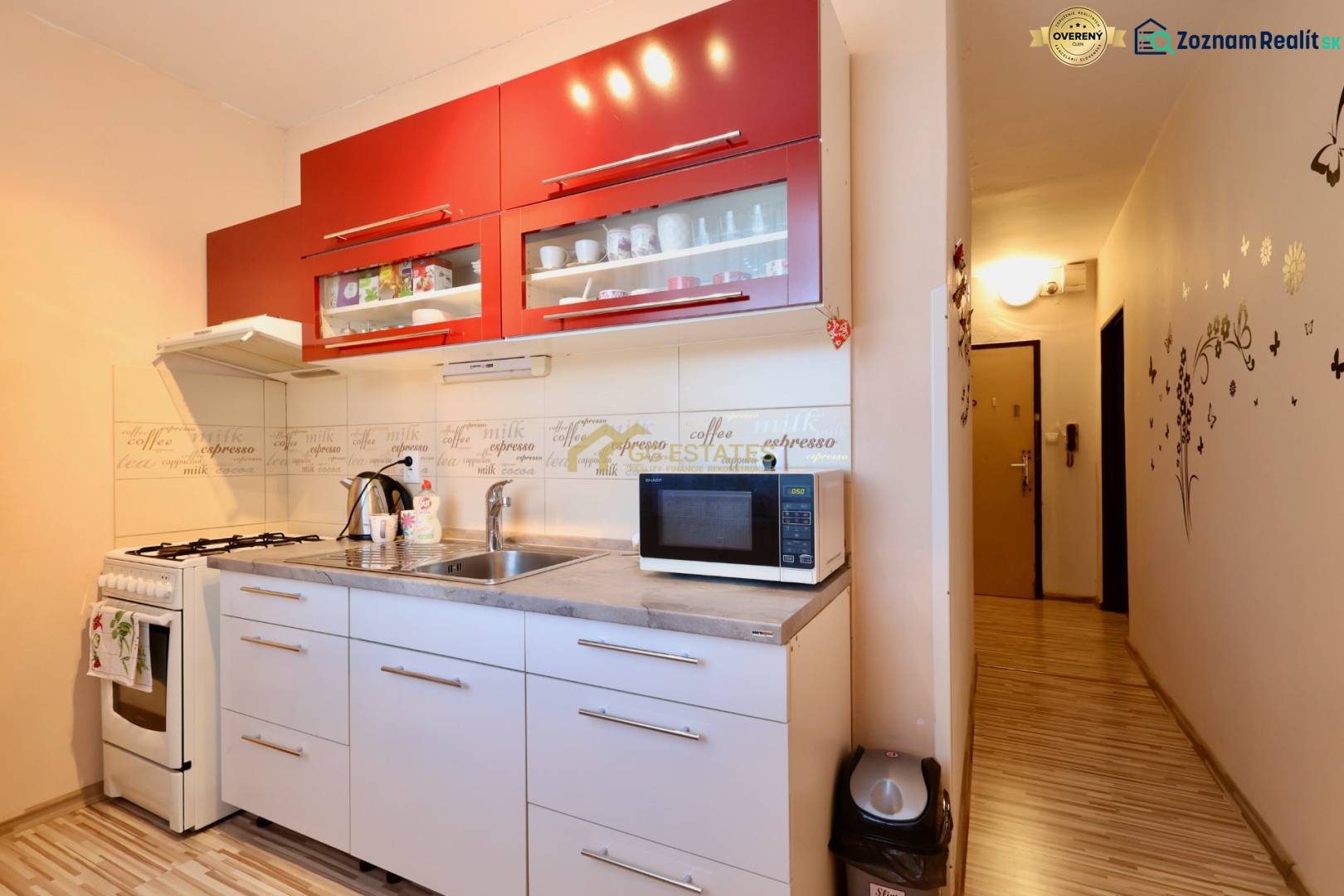 A kitchen space in a studio apartment with a white kitchen unit, red cabinets, and a wooden-patterned floor.
