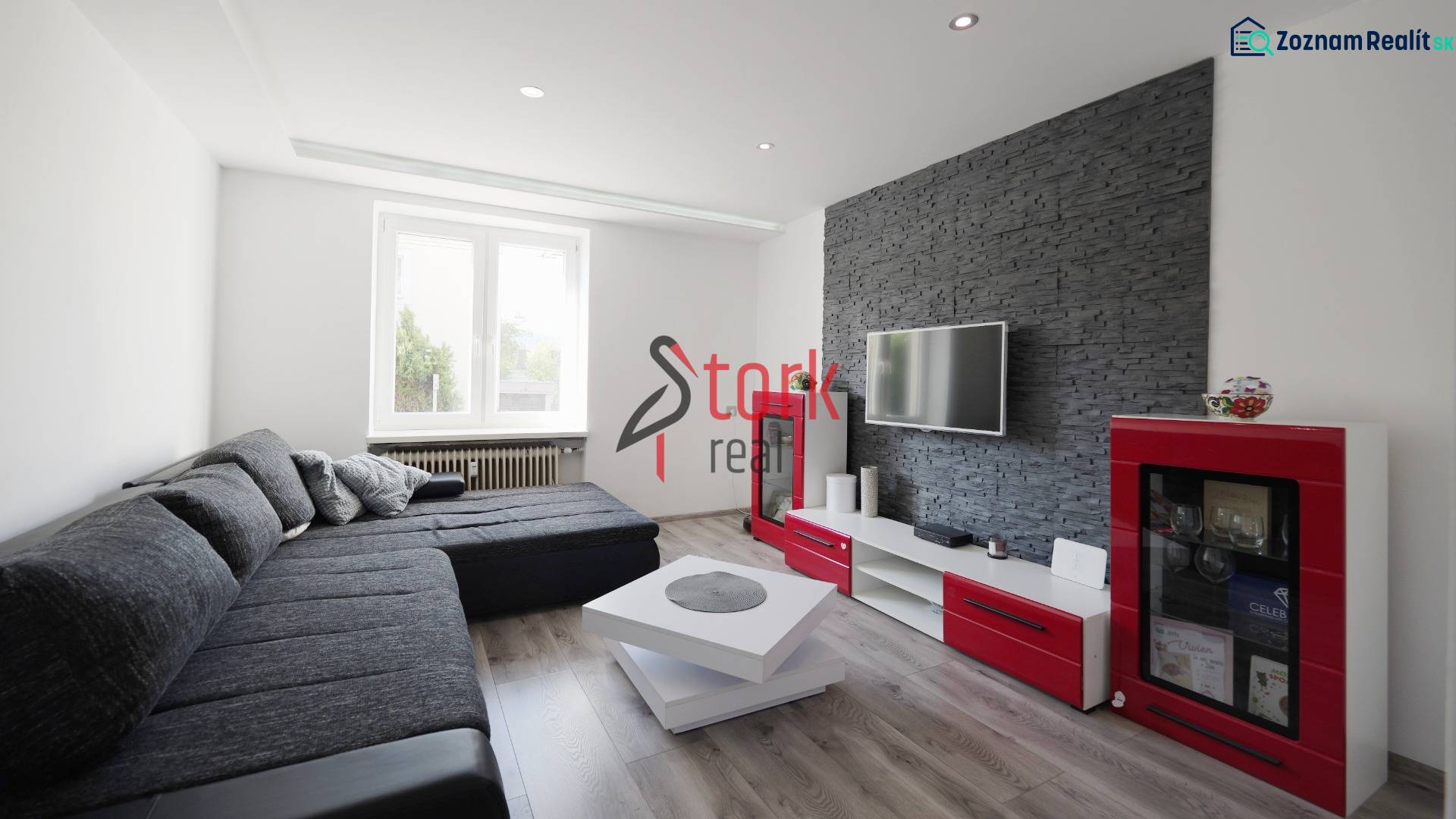 Living room in a two-room apartment with a dark gray sofa, television, and wooden decor flooring.