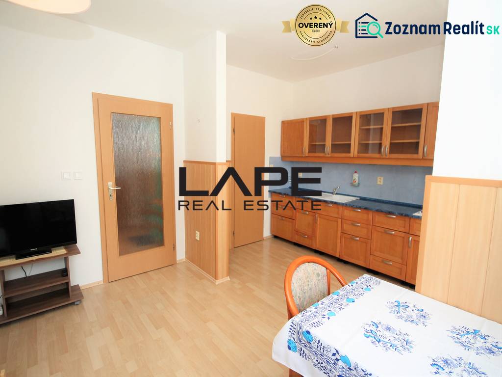 A kitchen in a studio apartment with a wood-patterned floor, kitchen unit, and television.