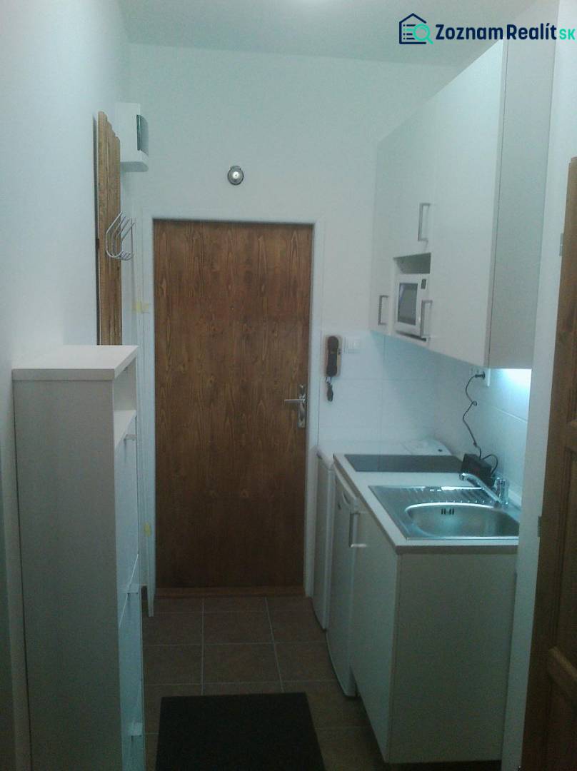 A kitchenette in a one-room apartment with white cabinets and wooden doors.