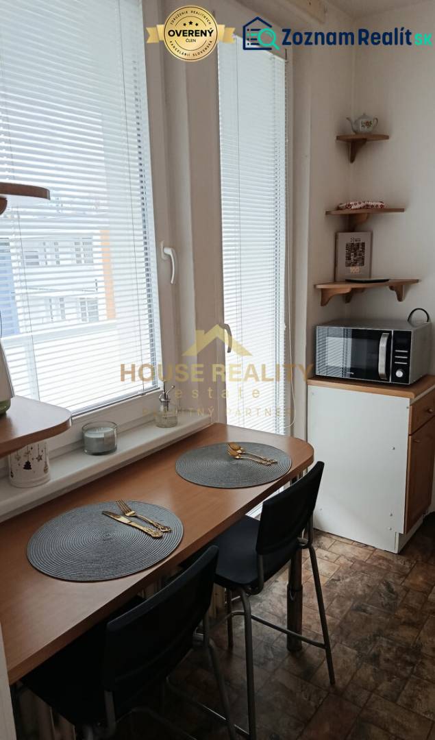 A kitchen with a dining area, a microwave, and windows with blinds in a studio apartment.