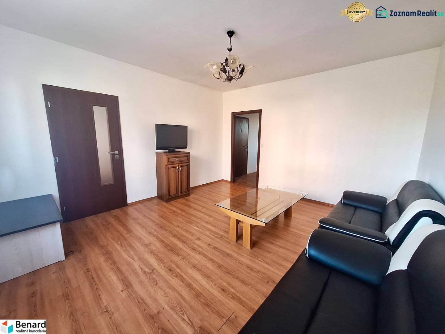 Living room in a three-room apartment with a wooden decor floor, black sofa, and television.