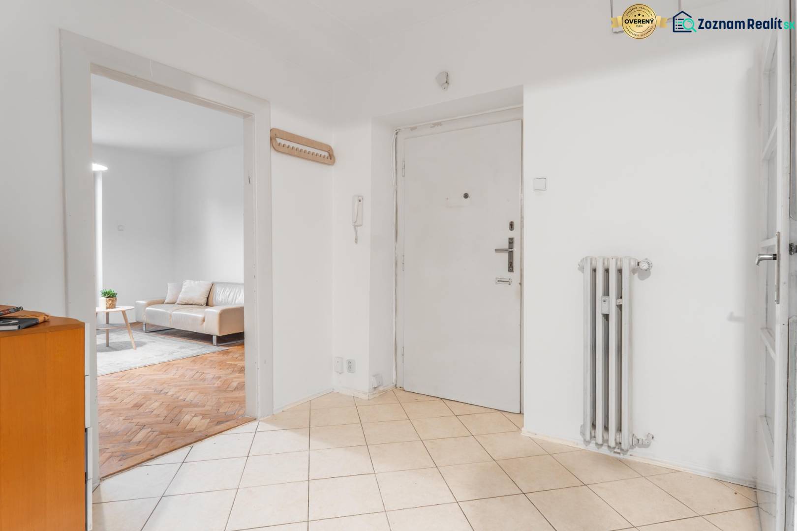 A hallway in a 3-room apartment with an entrance door, a radiator, and a view of the living room.