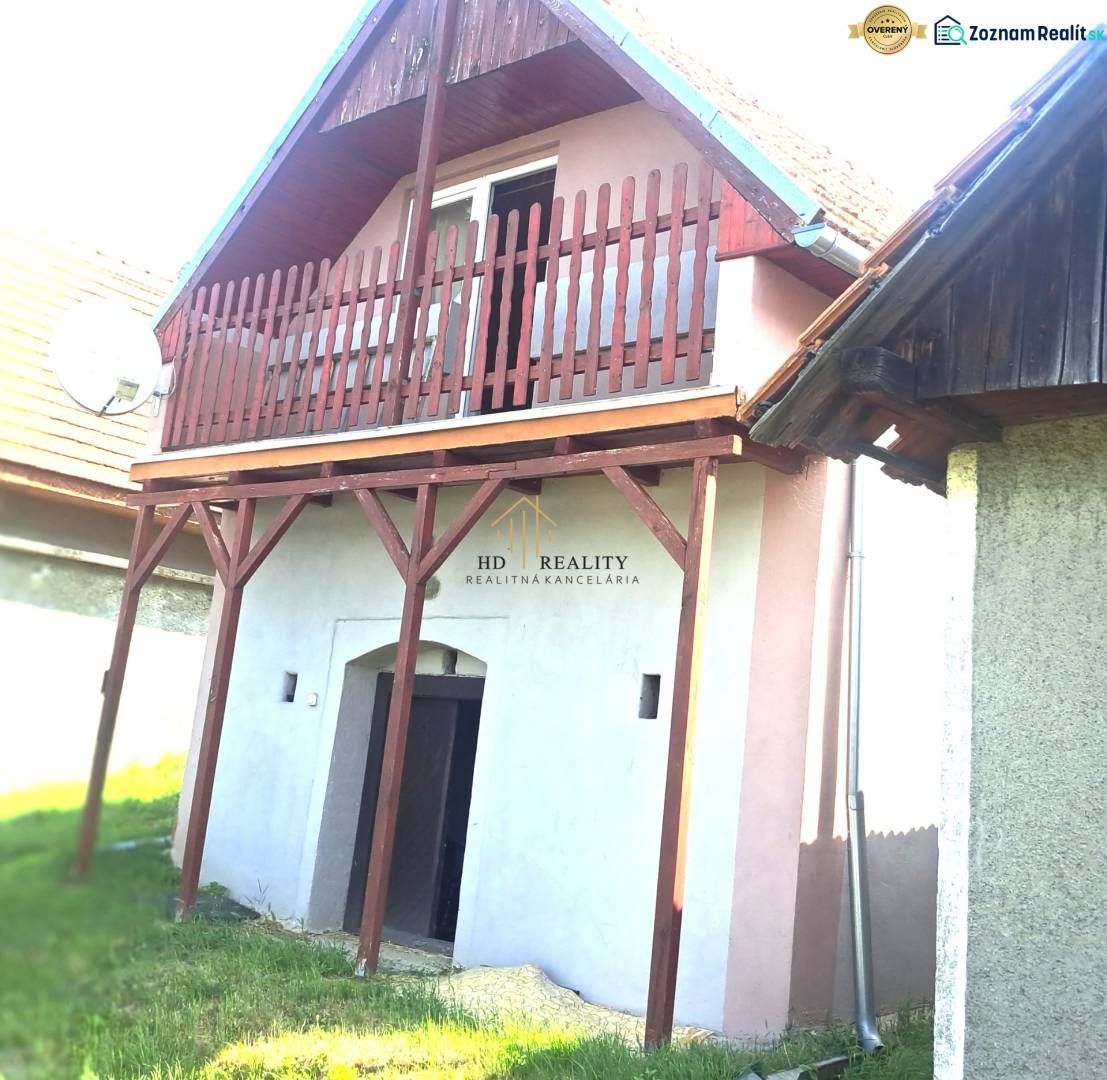 A red balcony on a cottage in Hontianske Trsťany, next to a green lawn and a neighboring house.