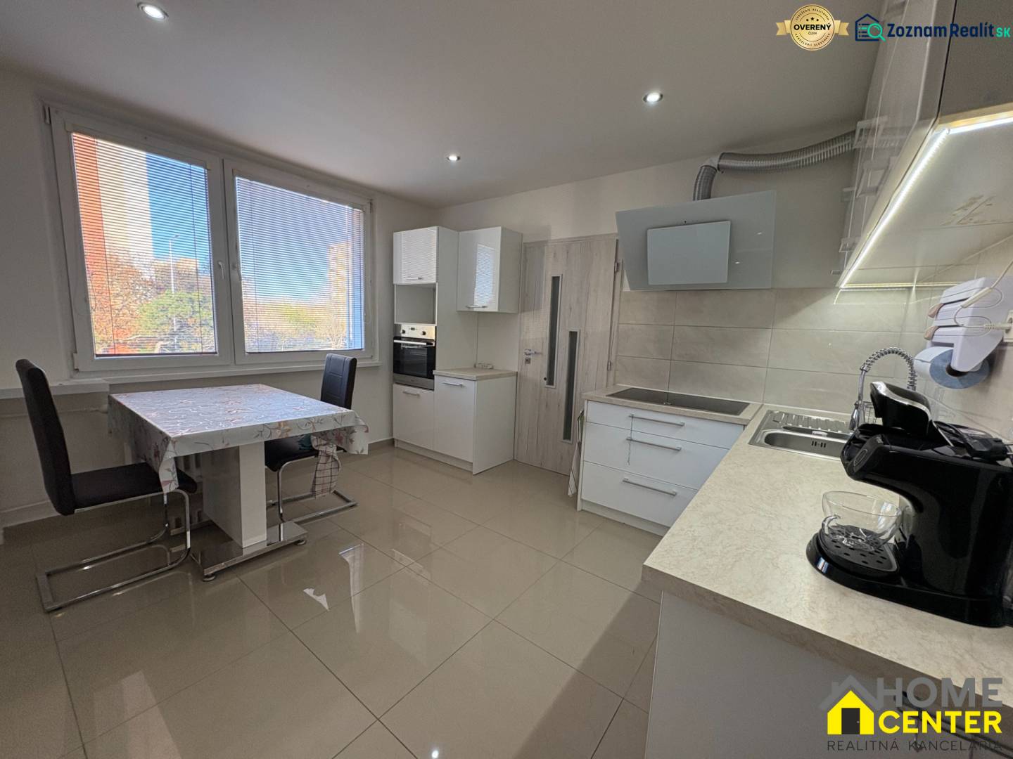 The kitchen of a 3-room apartment with large windows, a dining table, and light flooring.