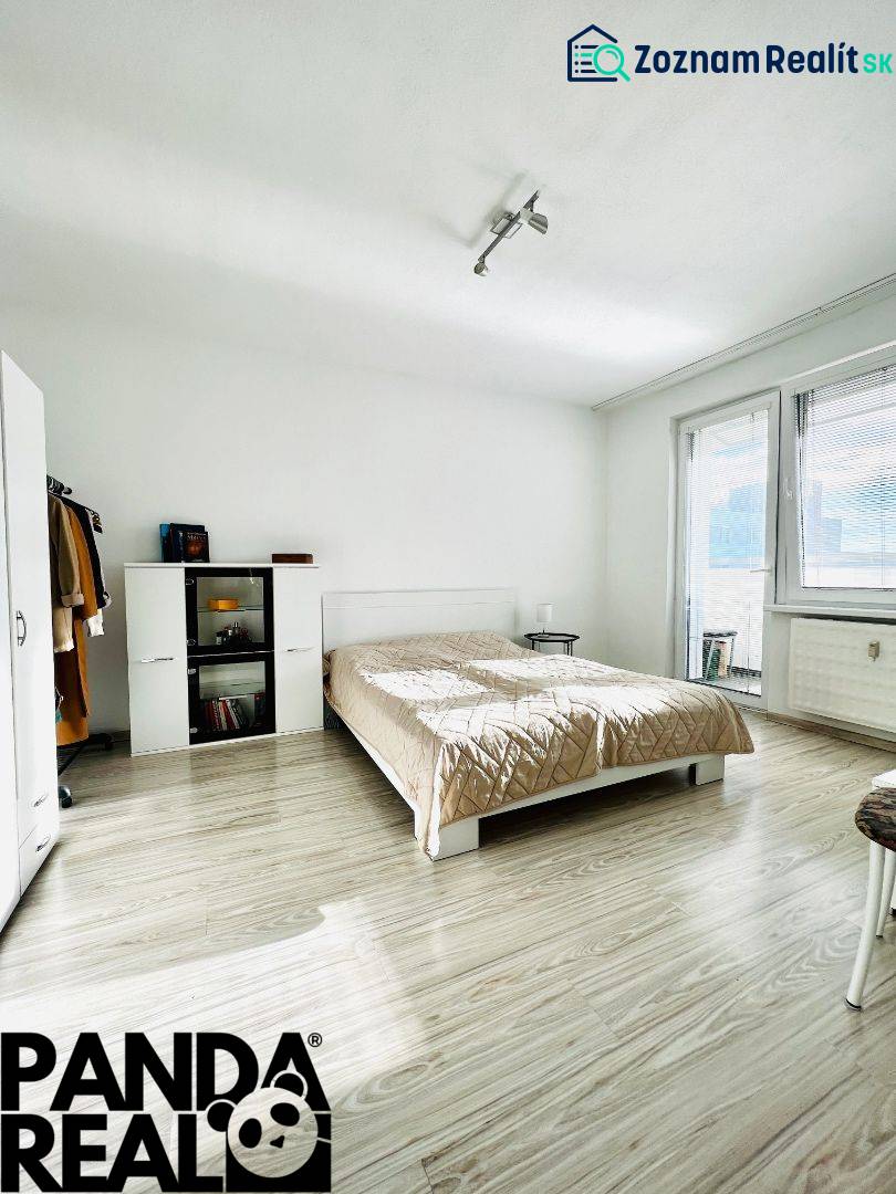 A bedroom in a studio apartment with a wood-patterned floor and light-colored furniture.