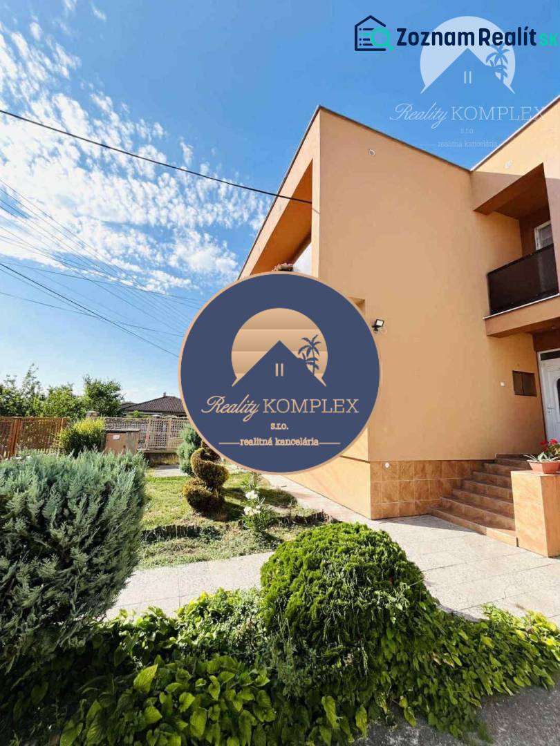 A family house in Okoč with a front garden and landscaped greenery in sunny weather.