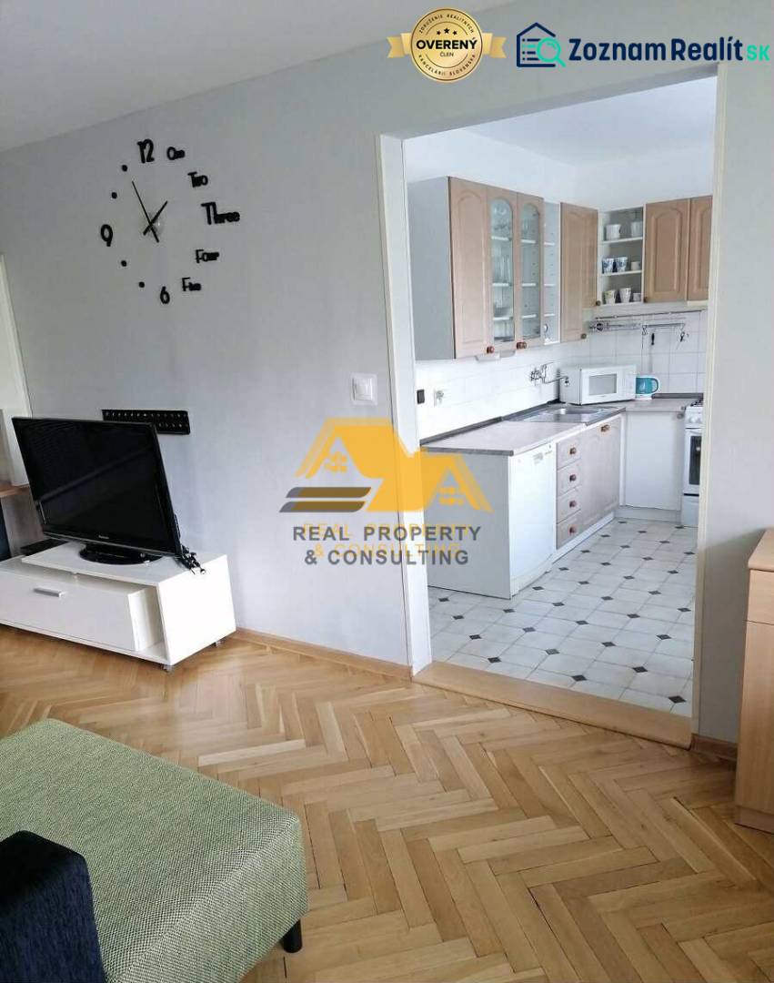 Living room in a three-room apartment with a wooden decor floor and a kitchen in the background.