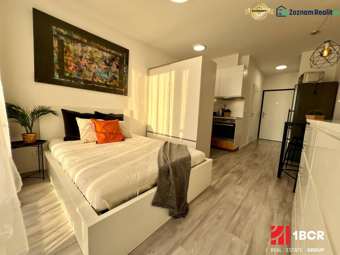 Interior of a studio apartment with a bed, kitchen, and wooden decor flooring.