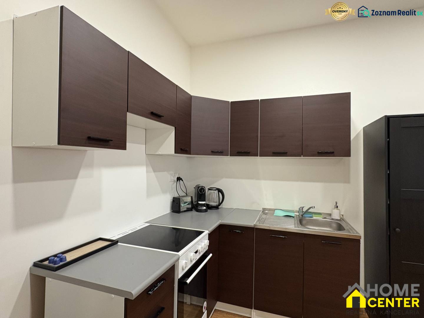 A kitchen in a studio apartment with dark cabinets and appliances, wooden decor flooring.