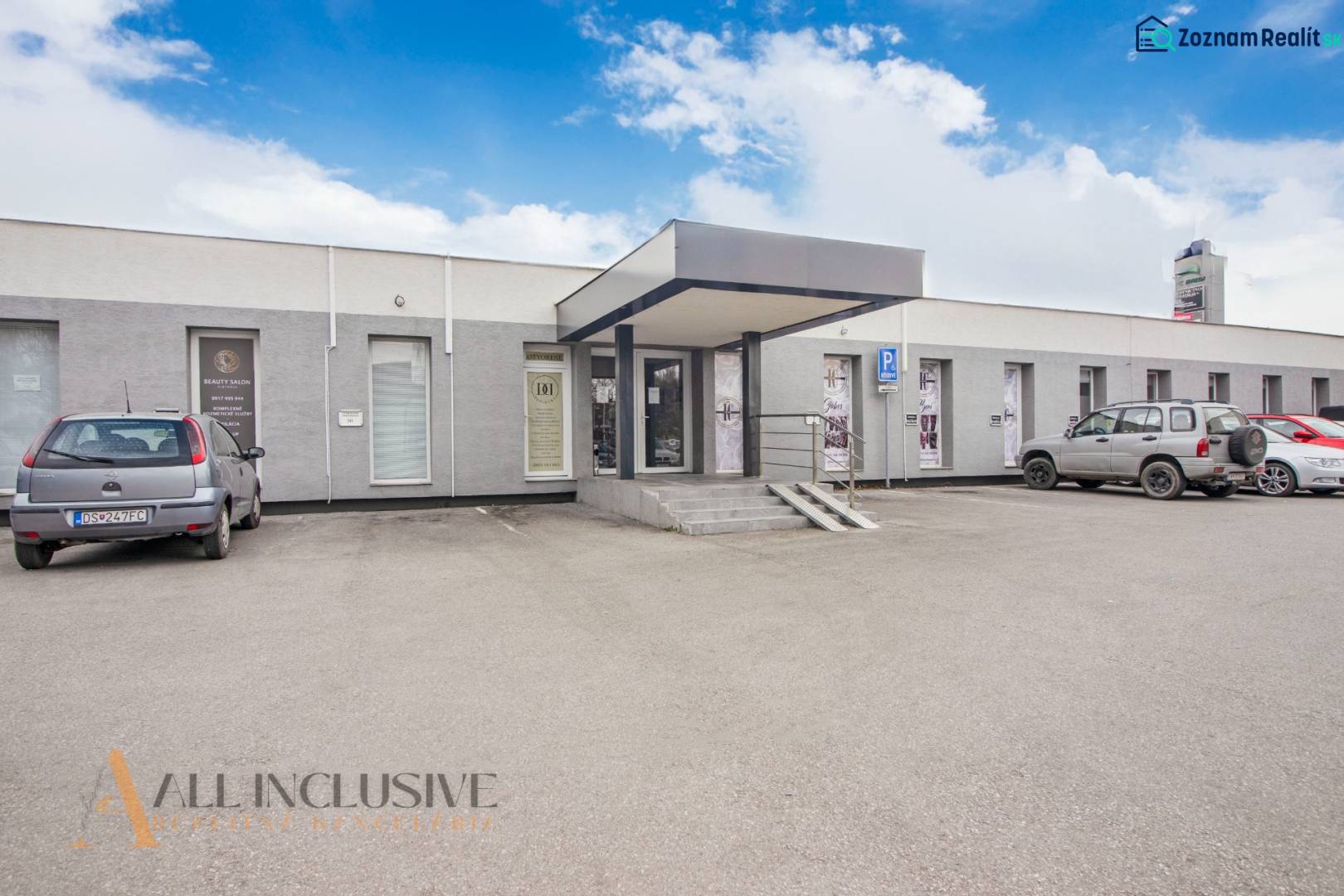 A single-story building with commercial spaces on Vajanského Street, a parking lot, and cars in front of the entrance.