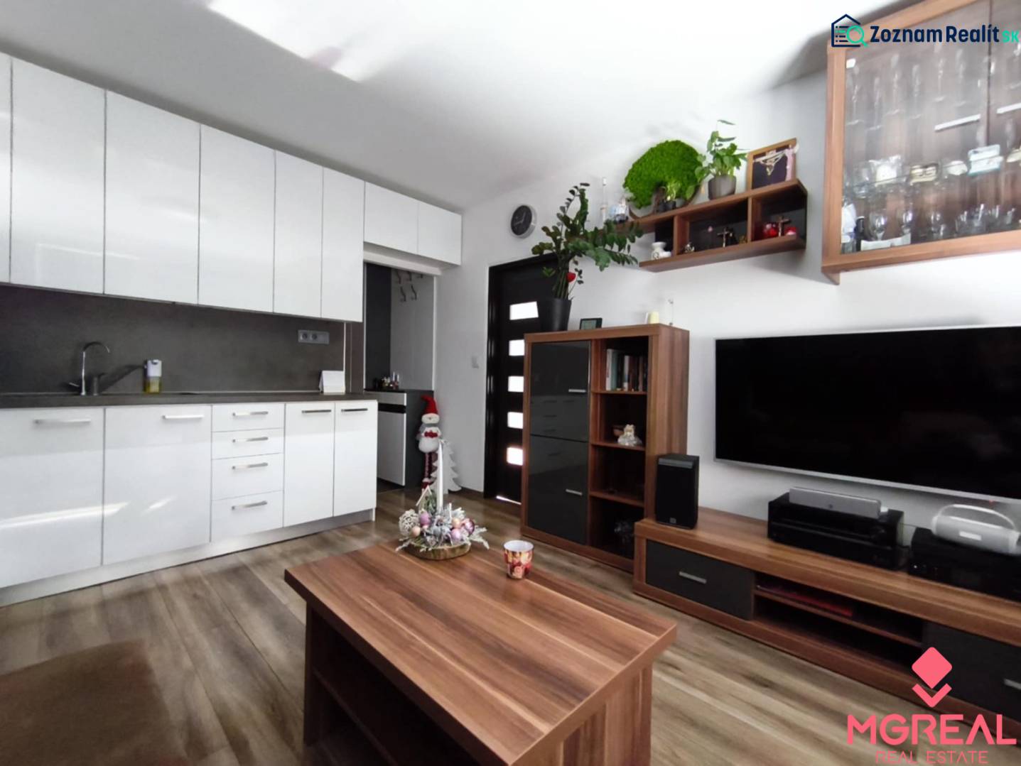 A kitchenette with white cabinets, a television, furniture, and a wooden decor floor in a studio apartment.