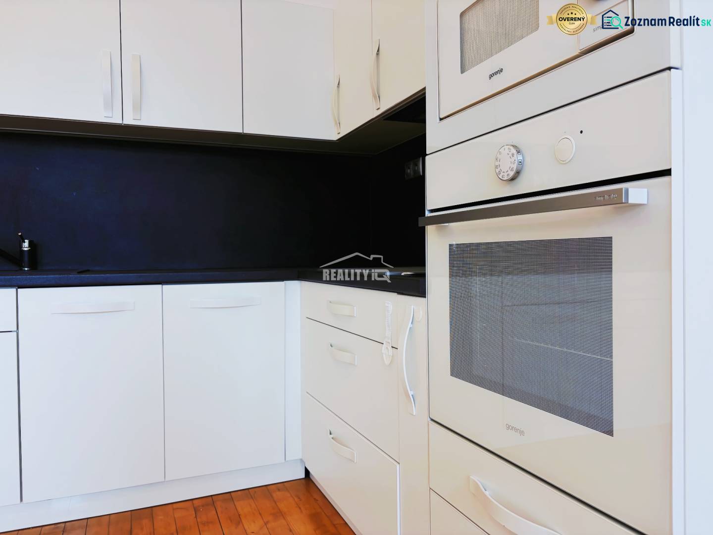 A kitchen in a 2-room apartment with white cabinets, a black countertop, and a wooden-patterned floor.