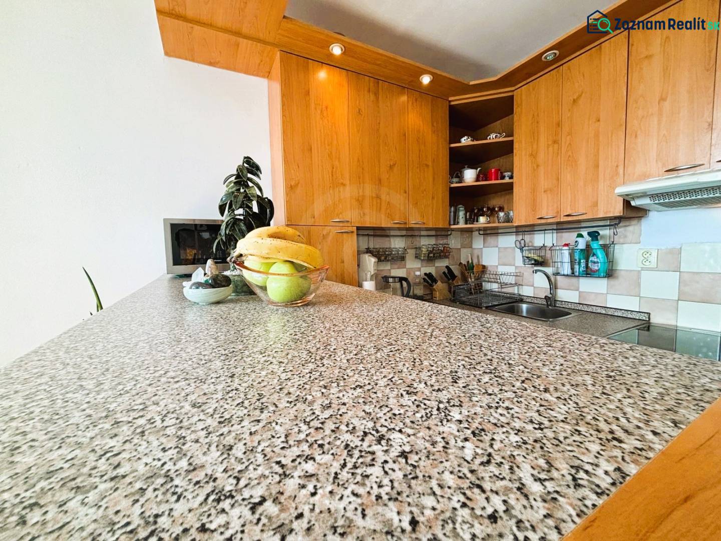 A kitchen in a 2-room apartment with a wooden decor and a fruit bowl on a stone countertop.