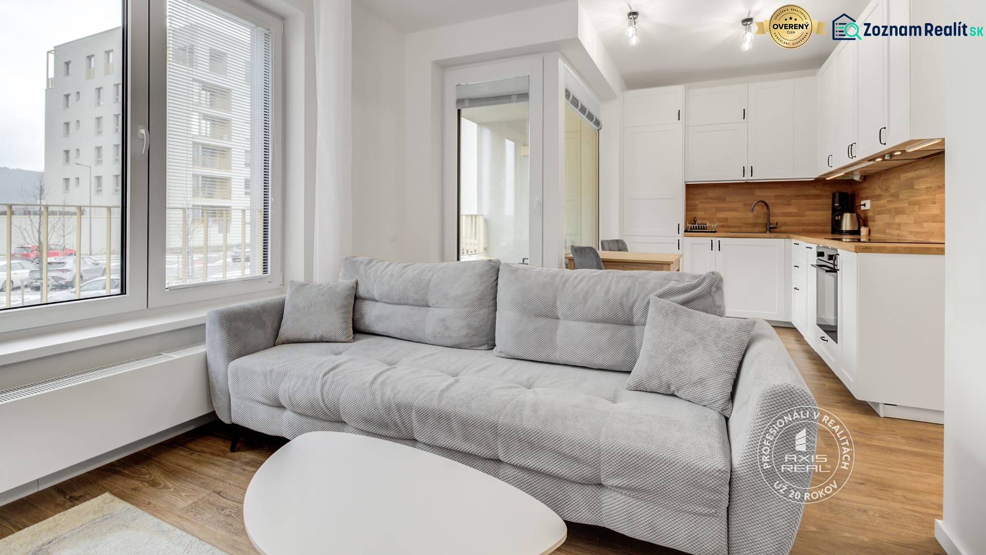 A bright living room with a kitchenette, a gray sofa, and a wooden decor floor in a studio apartment.