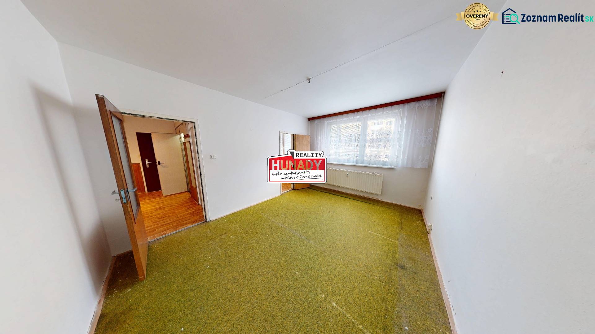 A room in a 3-room apartment with a carpet, a large window, and a door with a wooden decor floor.