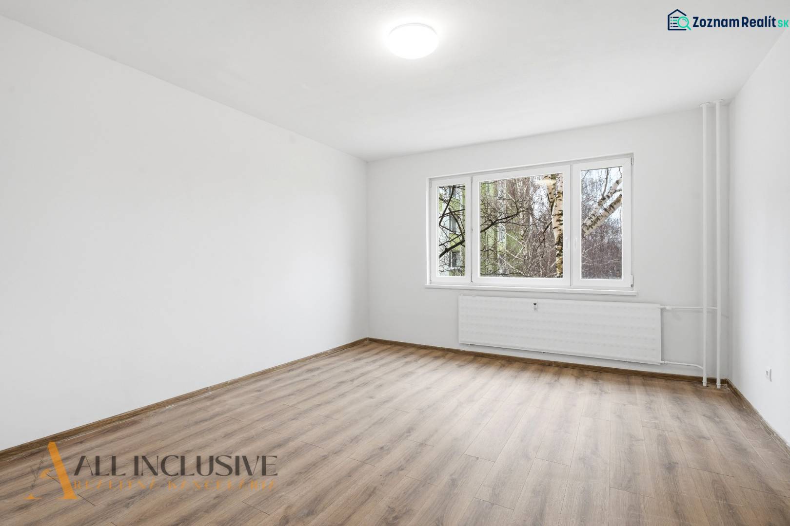 An empty living room in a three-room apartment with wood-patterned flooring and a large window.