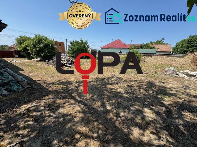 Building plot near a family house in Mužla with a grassy area, trees, and neighboring buildings.