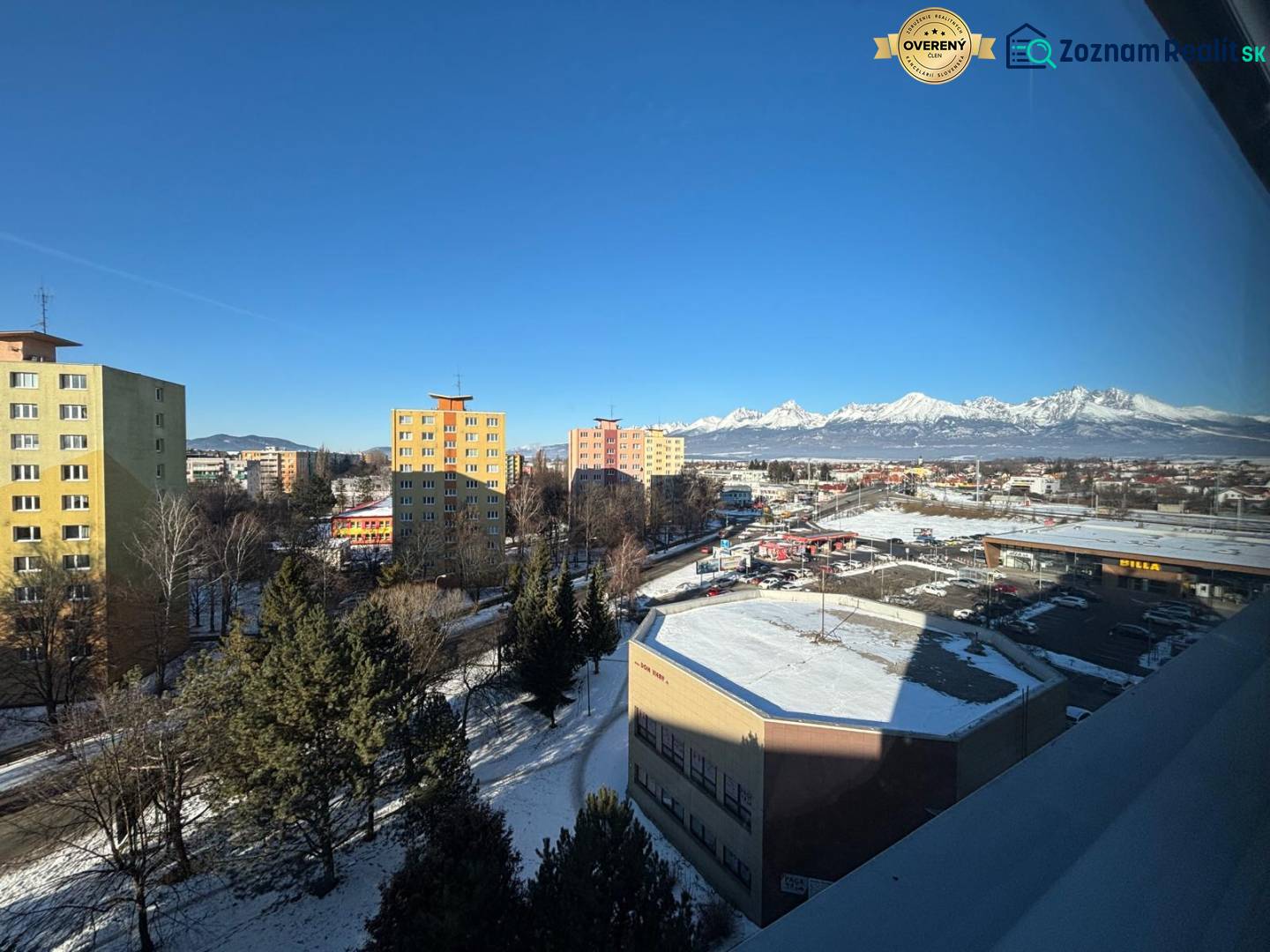 Poprad: apartment buildings, supermarkets, and snow-covered mountains behind offices under a clear sky.