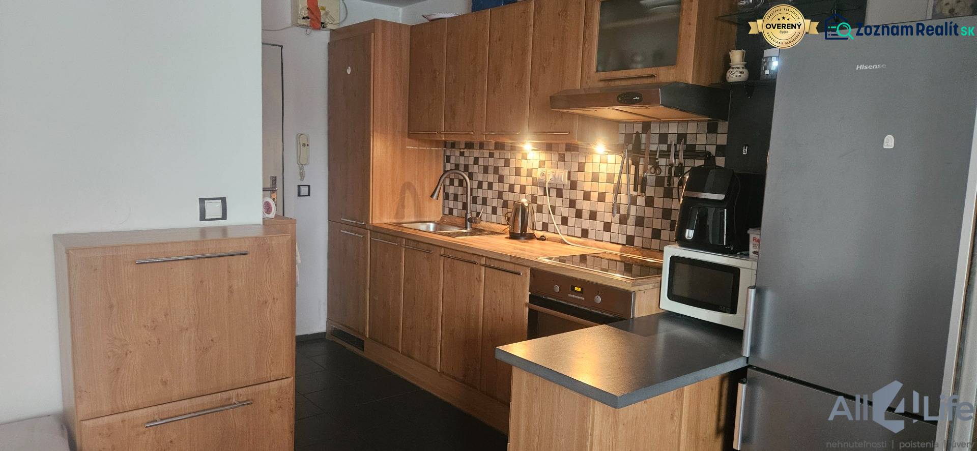 A kitchen in a studio apartment with wooden cabinets, tiled backsplash, appliances, and a white wall.