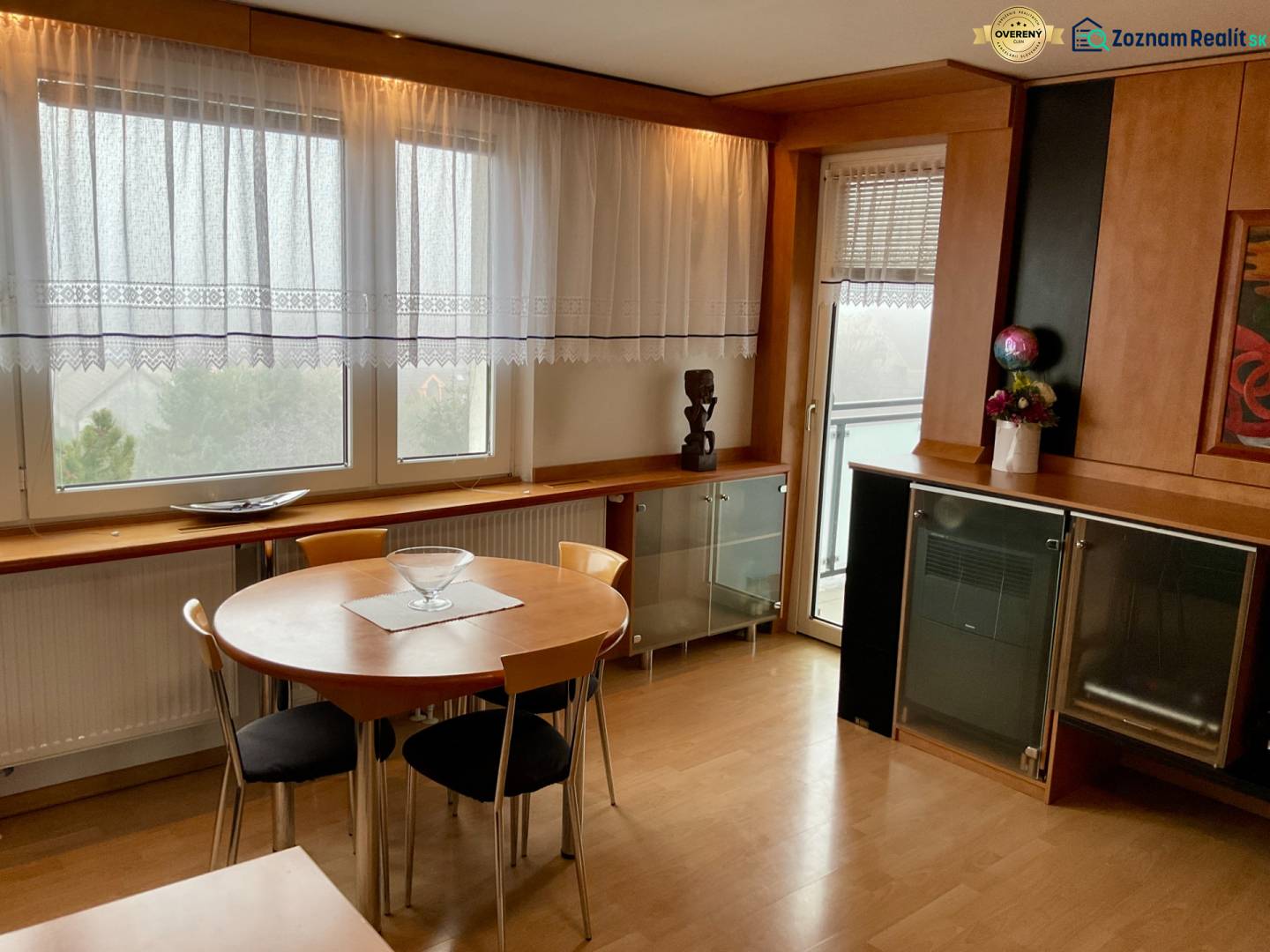 A dining room with wooden decor and windows in a 3-room apartment, overlooking greenery.