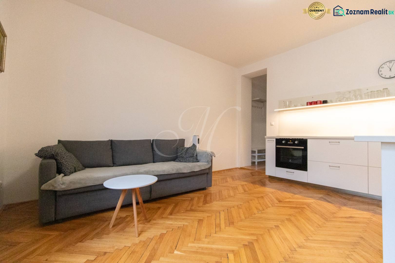 Living room in a two-room apartment with a sofa, kitchen unit, and wooden decor flooring.