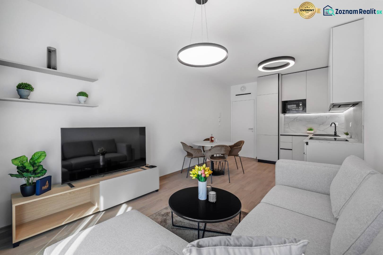 Living room with kitchen in a 2-room apartment, light couch, television, wooden decor flooring.