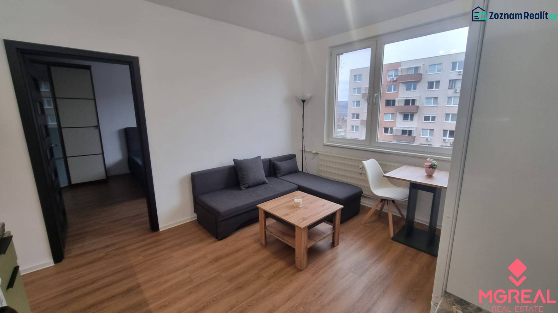 Living room in a studio apartment with a sofa, table, chair, and wood-patterned flooring.