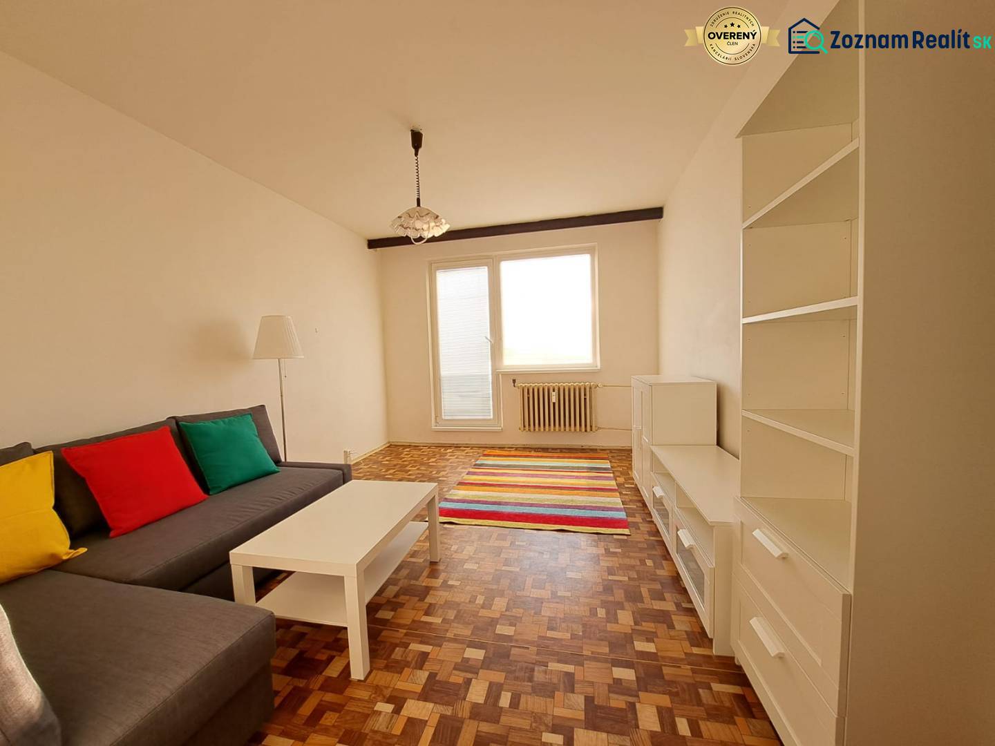 Living room in a two-room apartment with a sofa, white furniture, colorful pillows, and a wooden-patterned floor.