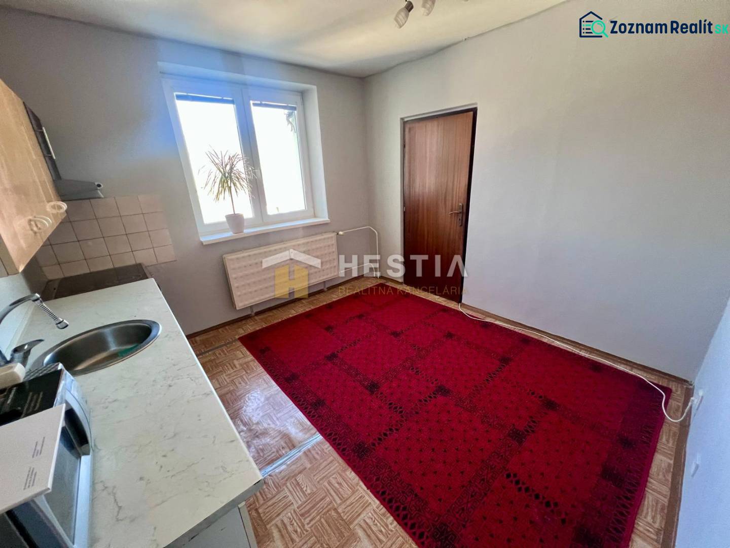 The kitchen of a three-room apartment with carpet, wooden decor flooring, and simple furnishings.
