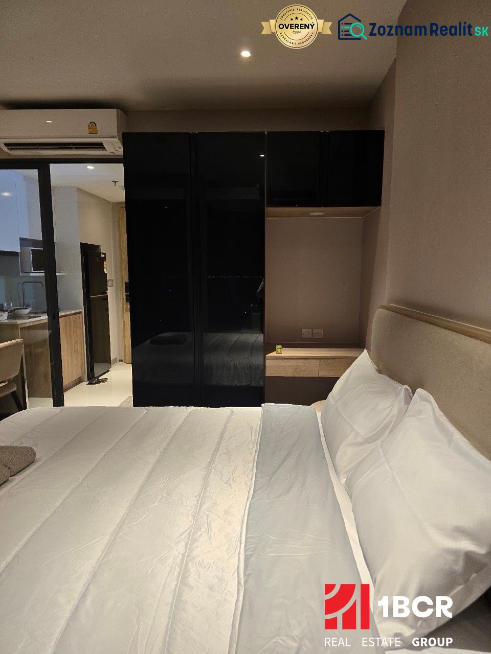 A bedroom with a white bed and a black wardrobe, next to the kitchen unit.