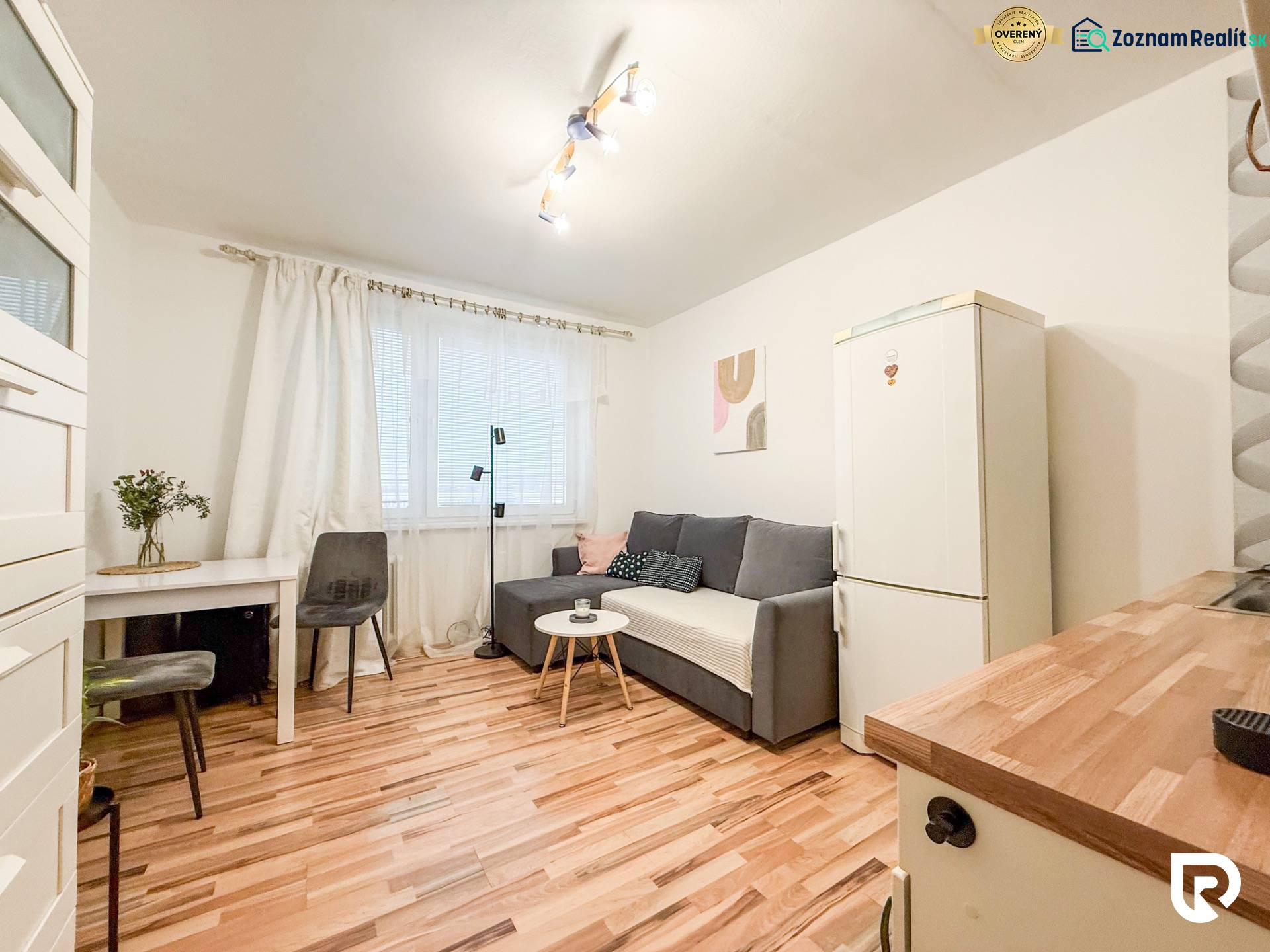 Studio apartment with wood-patterned flooring, gray sofa, and white refrigerator.