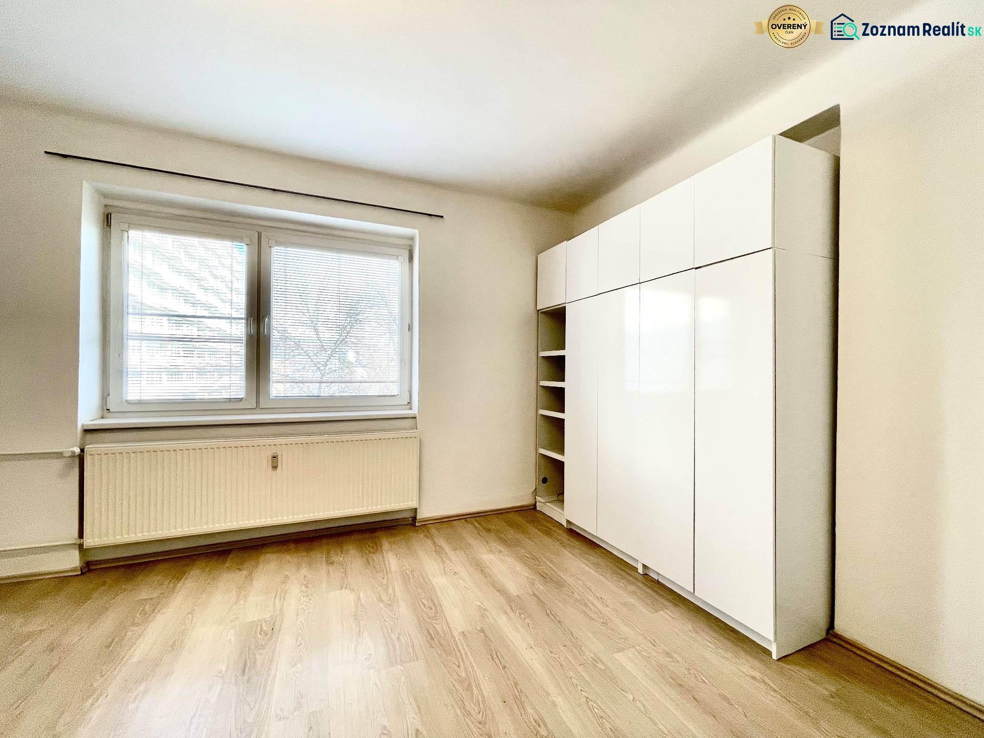 A room with a large window, a wardrobe, and a floor with a wooden decor in a studio apartment.