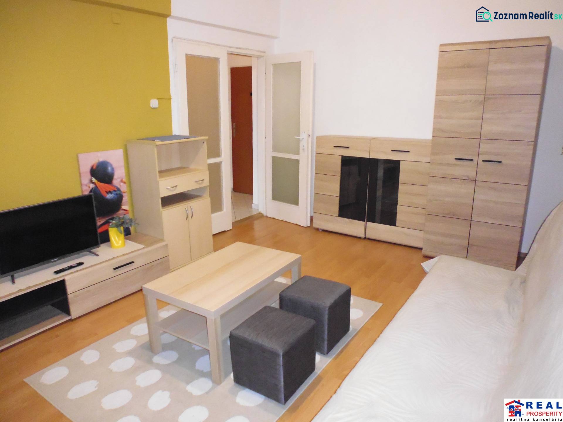 Living room in a studio apartment with flooring and wooden decor, furniture, and a television.