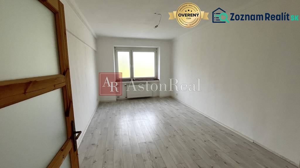 An empty room in a three-room apartment with a wood-patterned floor and a large window.