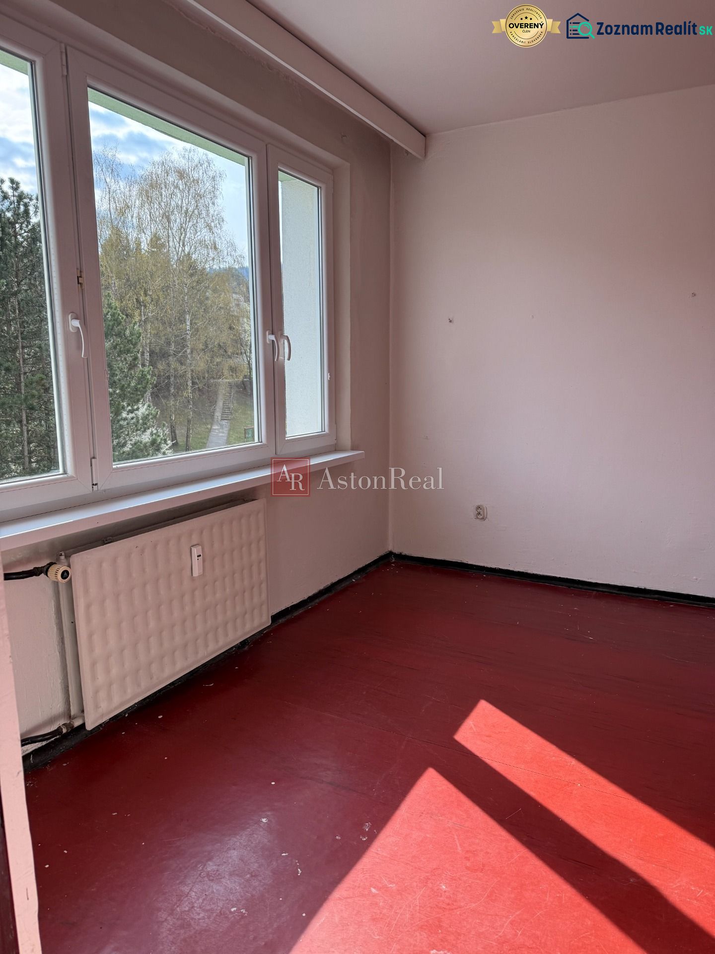 A room with a red floor in a 2-room apartment, a large window lets in natural light.