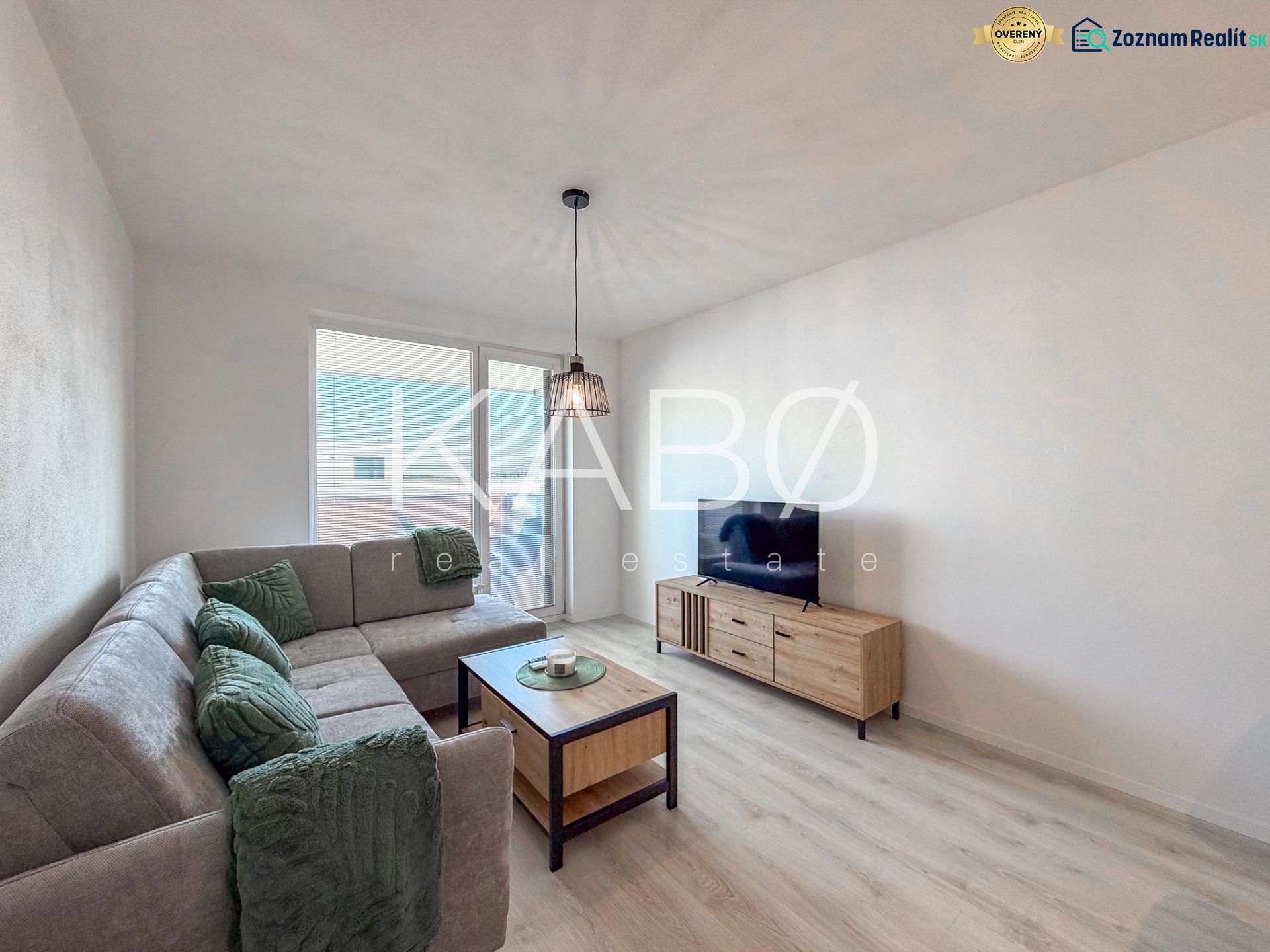 Living room in a two-room apartment with a large couch, television, and wooden-patterned flooring.