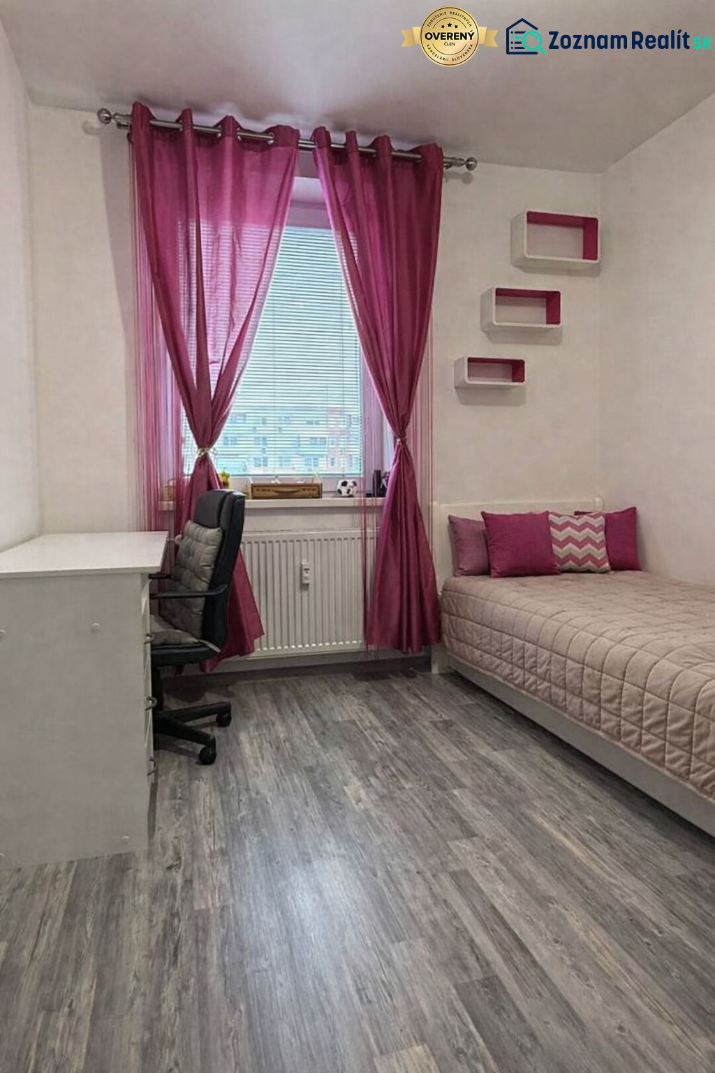 A room in a 4-room apartment with pink curtains, a bed, a table, and a wooden decor floor.