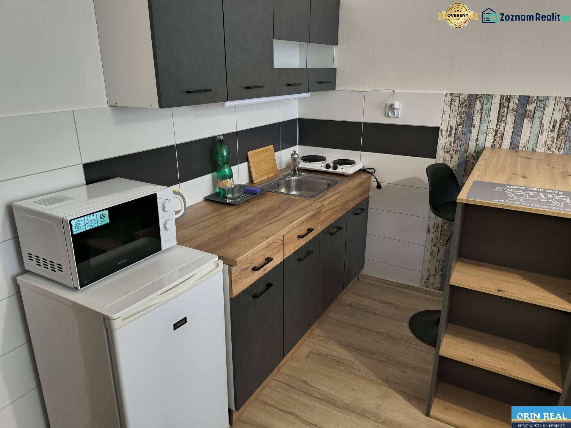 Kitchen area of a studio apartment with appliances and flooring with a wood decor.