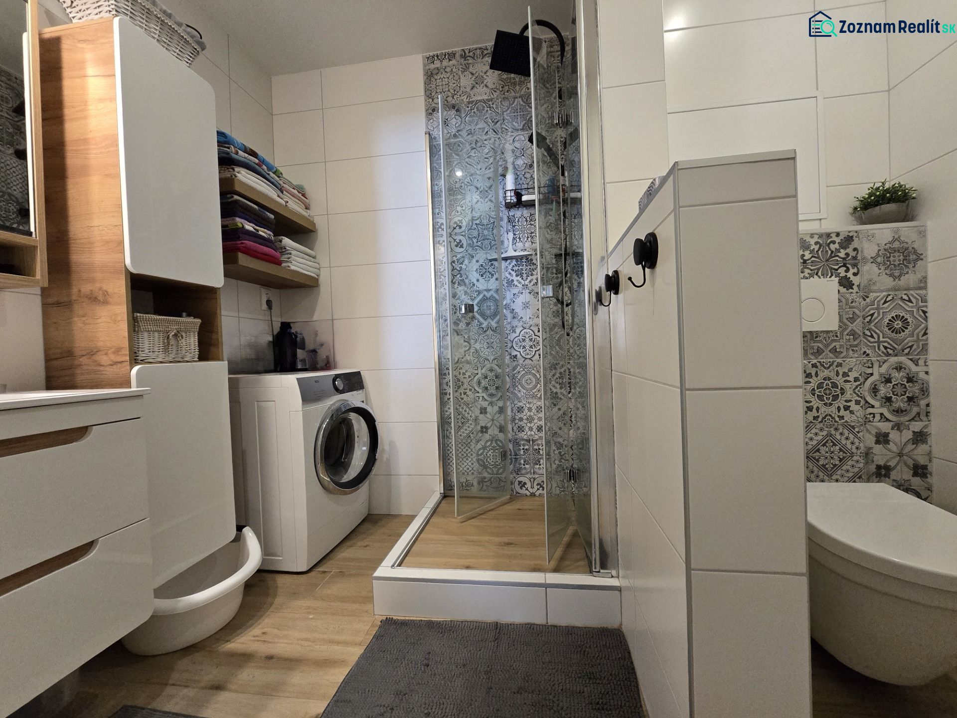 A bathroom in a 2-room apartment with a washing machine, shower, and wooden decor flooring.