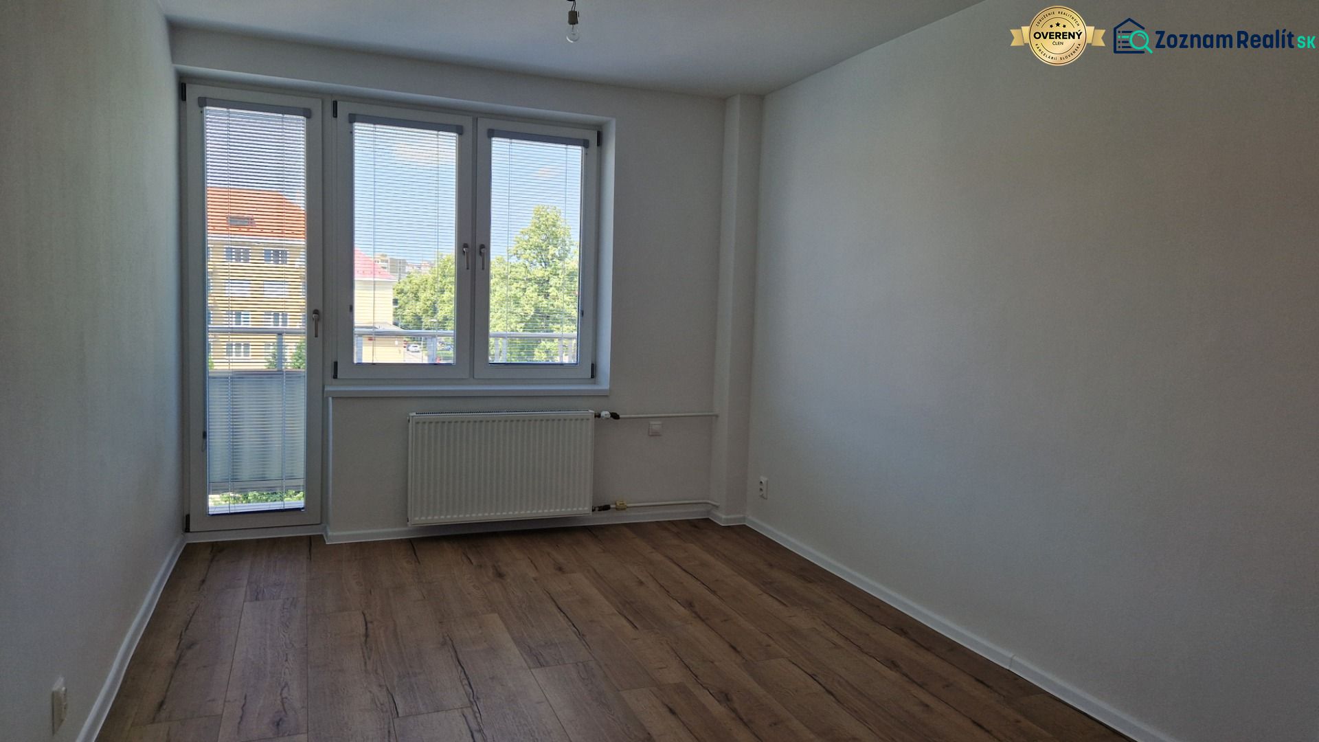 A room in a 3-room apartment with a wooden decor floor, a large window, and a radiator.