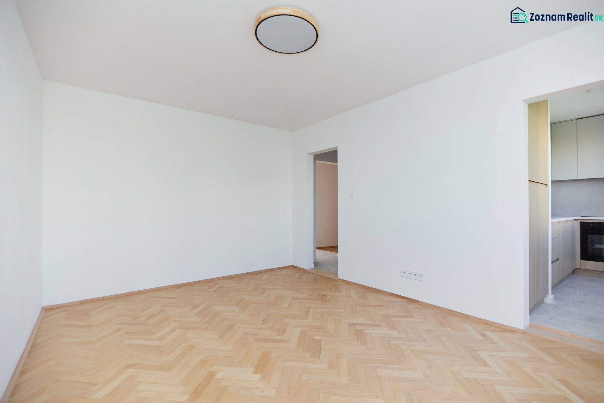 An empty room with wooden floor decor and a view into the kitchen in a 3-room apartment.