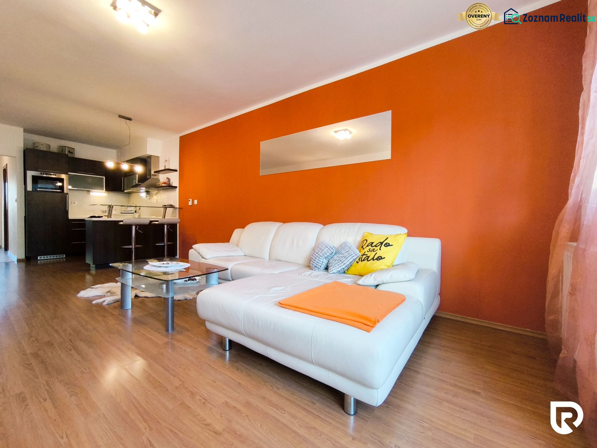 Living room of a two-room apartment with an orange wall, white sofa, and kitchen, floor with wooden decor.