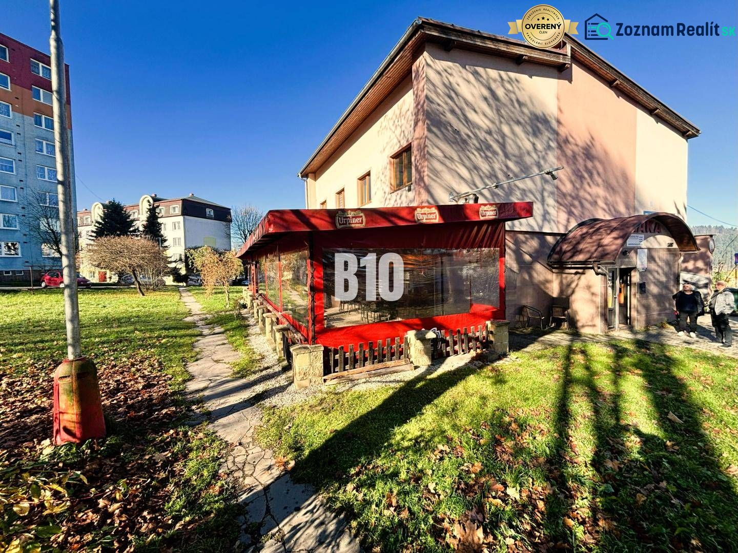 A building in Čadca with a garden, terrace, and red awning in sunny weather.