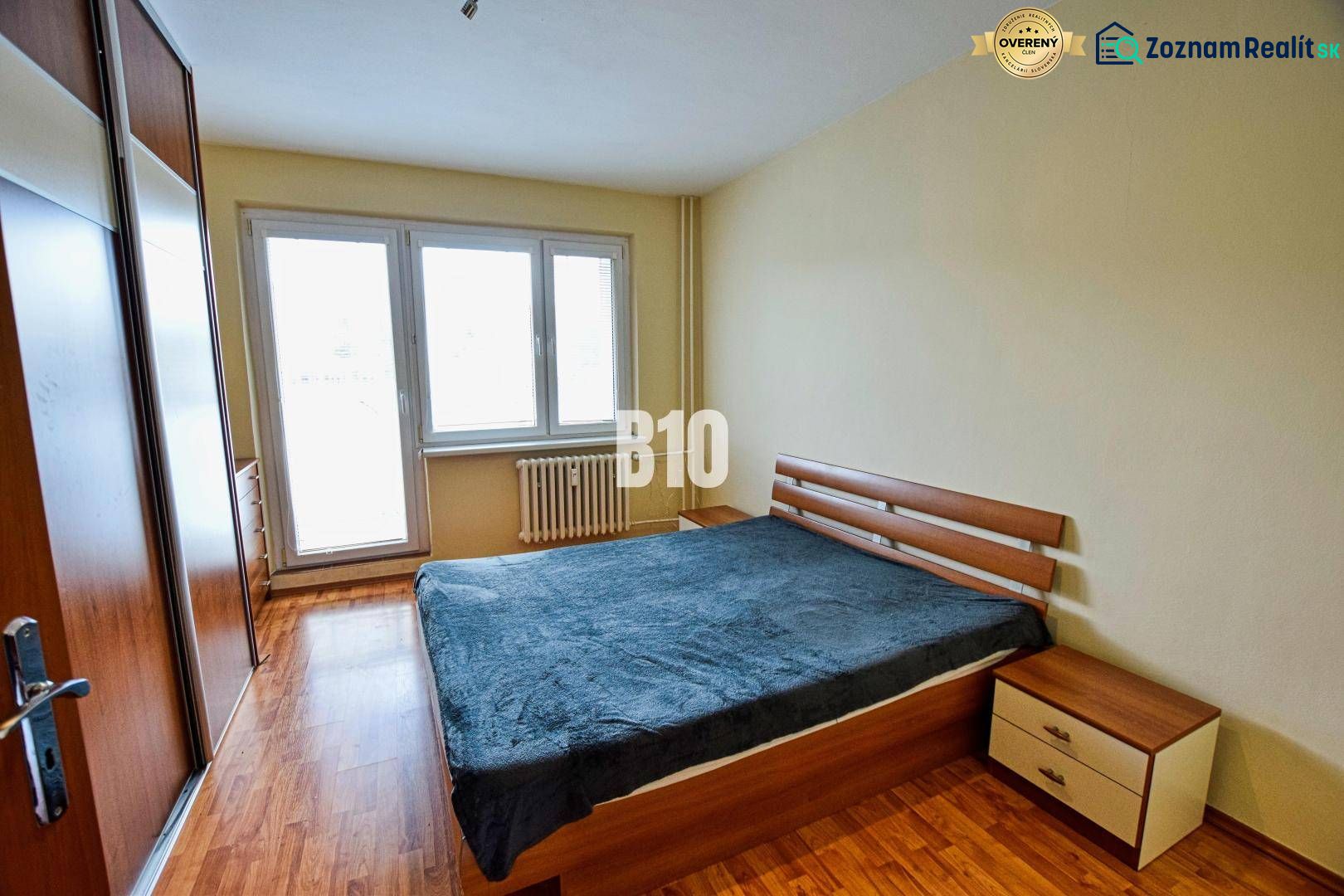 A bedroom in a 3-room apartment with a bed, wood-patterned flooring, and a built-in wardrobe.