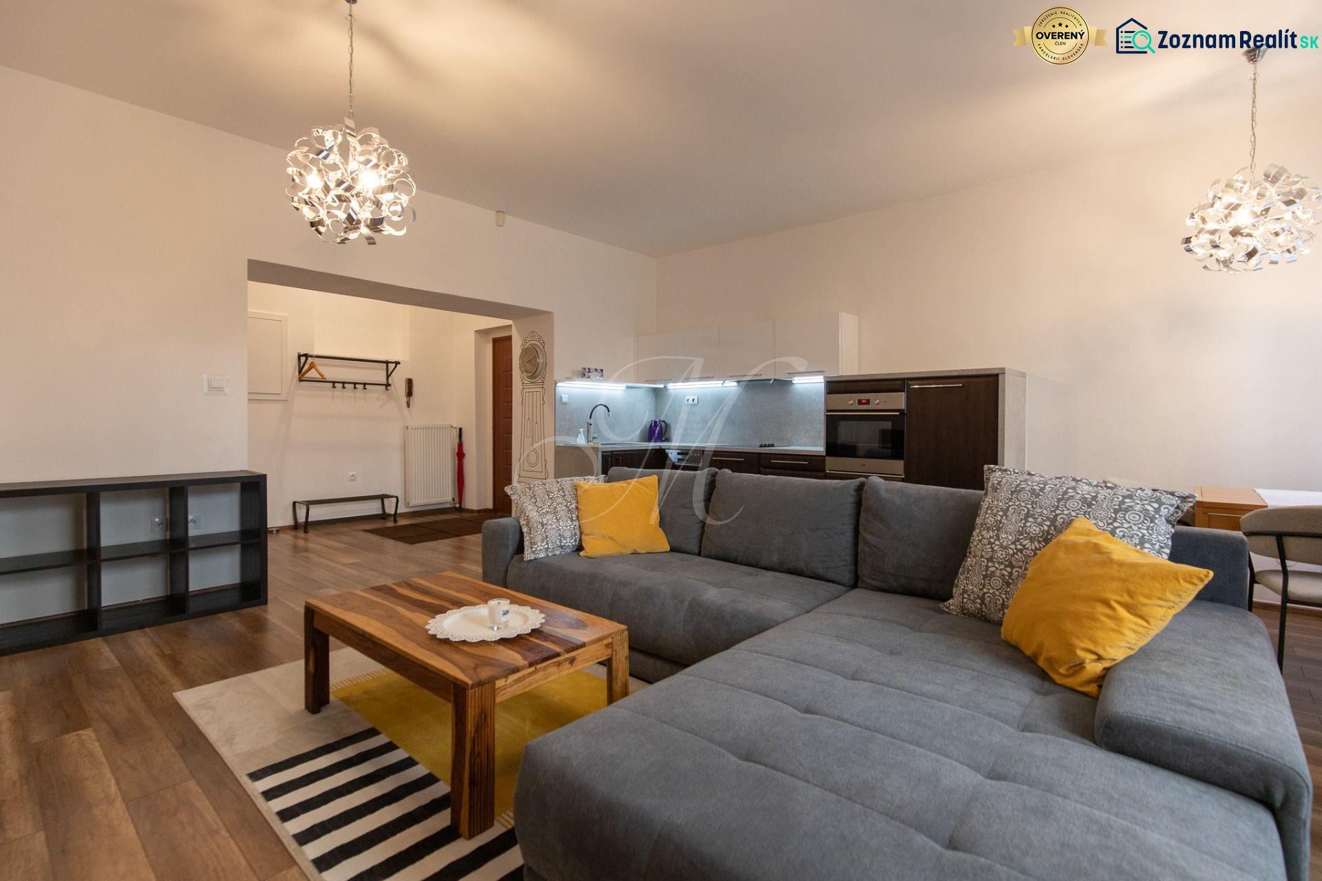 Living room with a kitchen, gray sofa, wooden table, and floor with a wooden design.