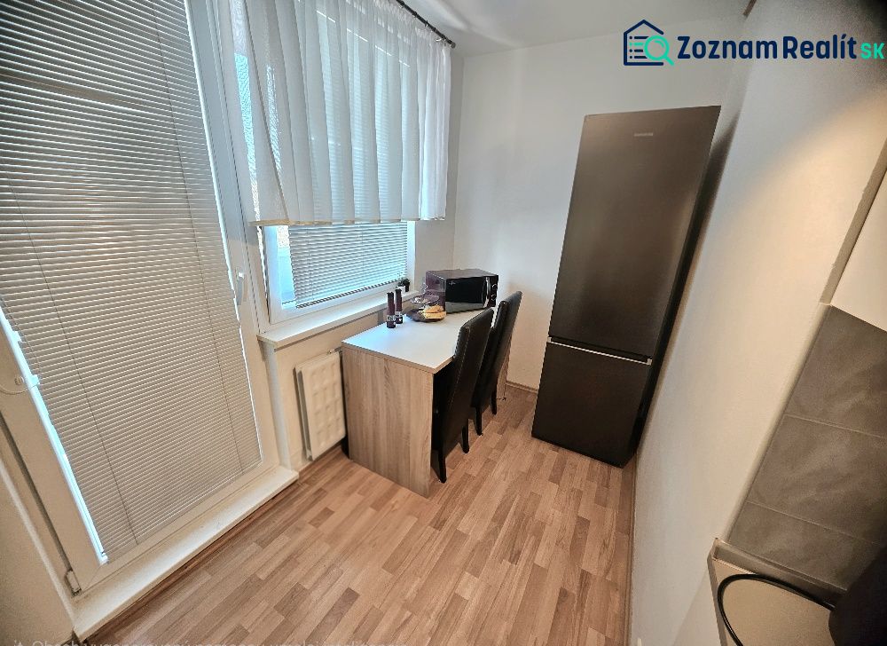 A kitchen in a studio apartment with a wood-patterned floor, a refrigerator, and a table by the window.