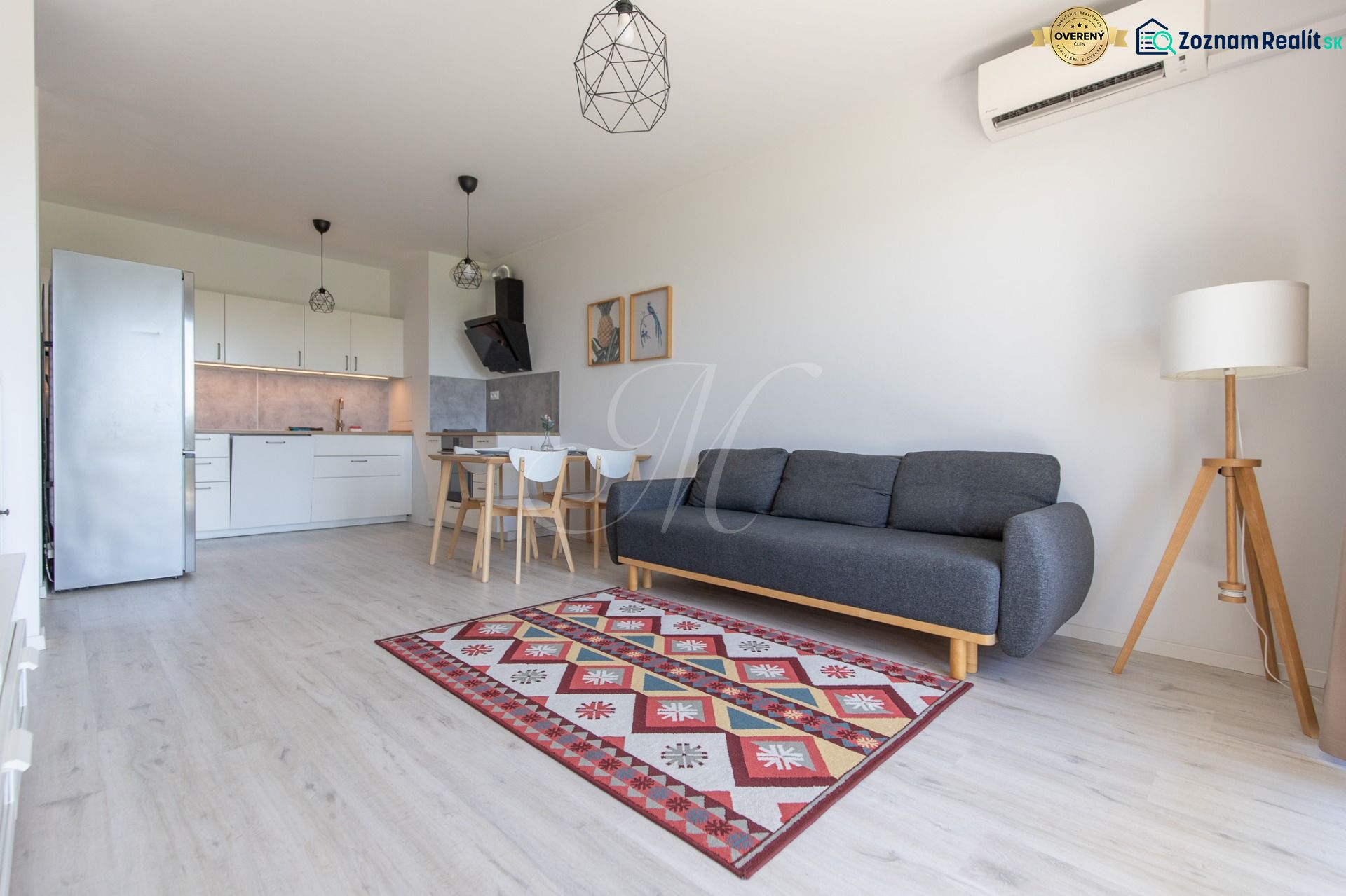 Living room with kitchen, gray sofa, wooden decor flooring, colorful carpet, dining table.