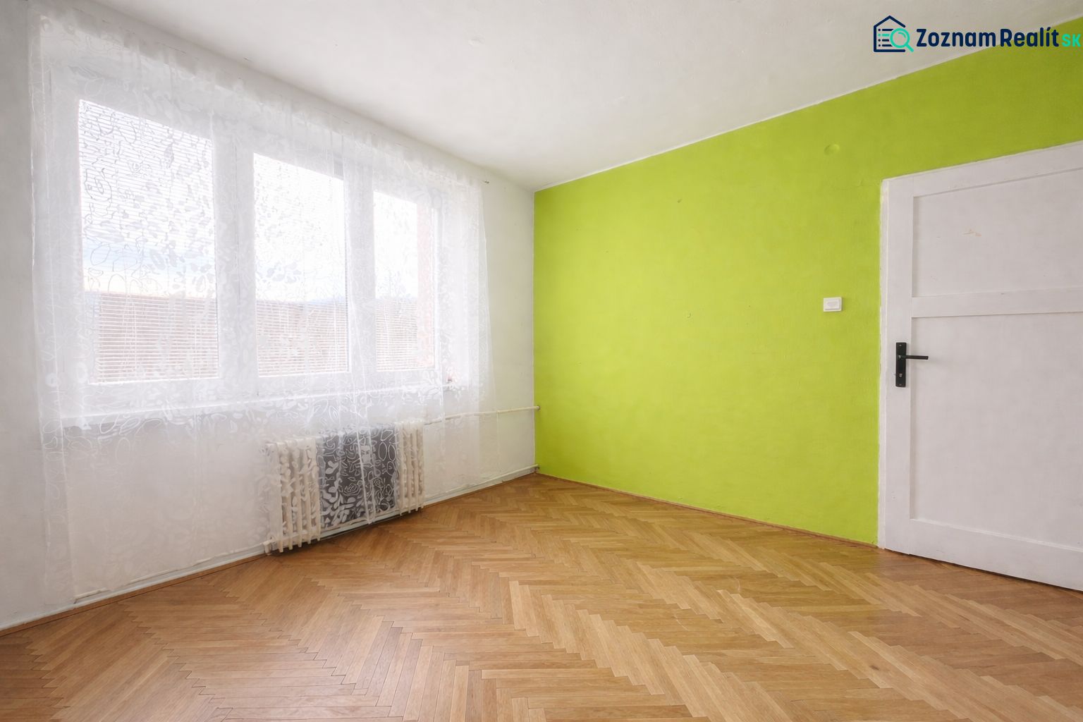 A room with a green wall, white curtains, and a wood-patterned floor in a two-room apartment.