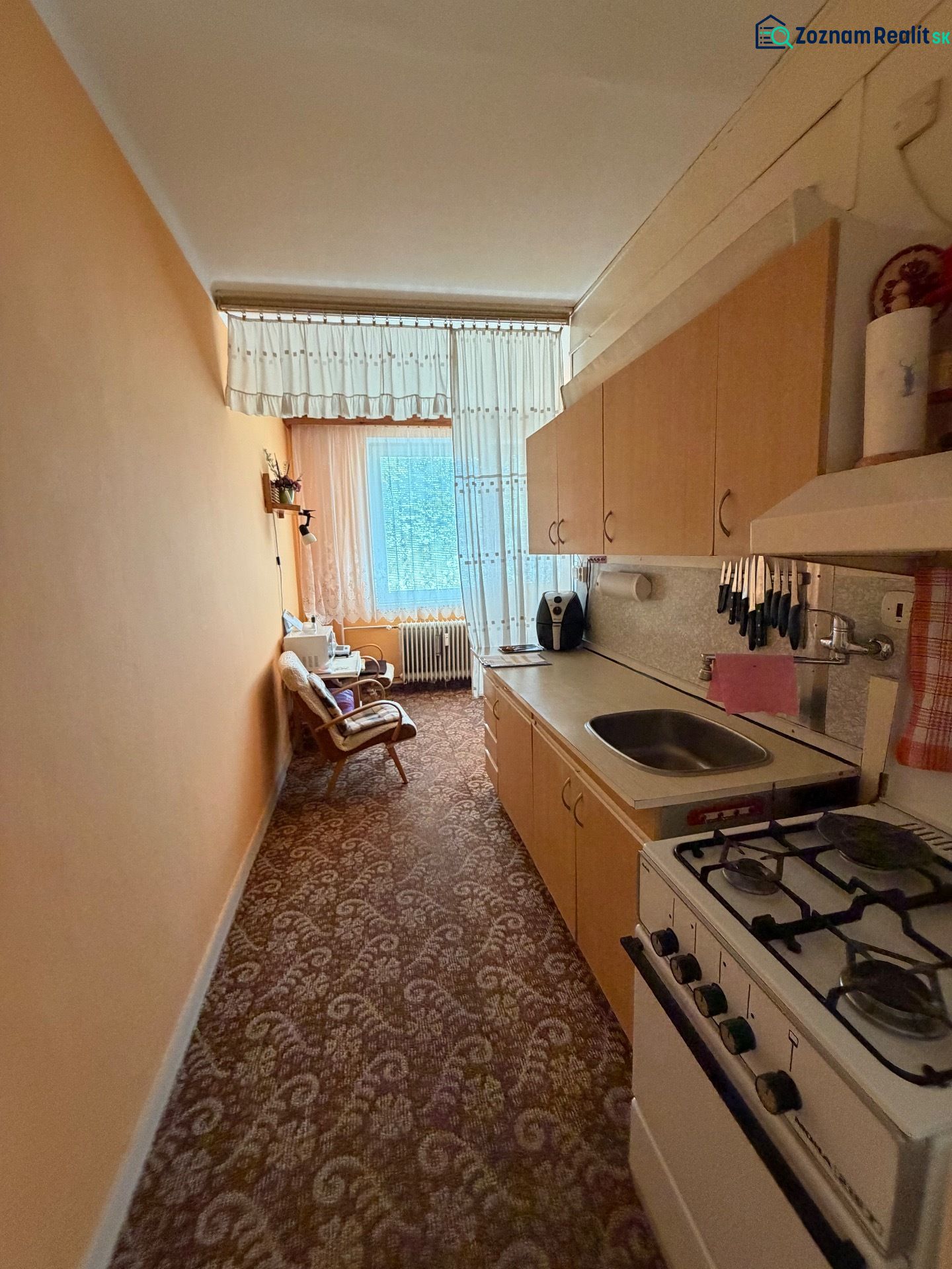 A narrow kitchen in a two-room apartment with a gas stove and a floral pattern on the carpet.